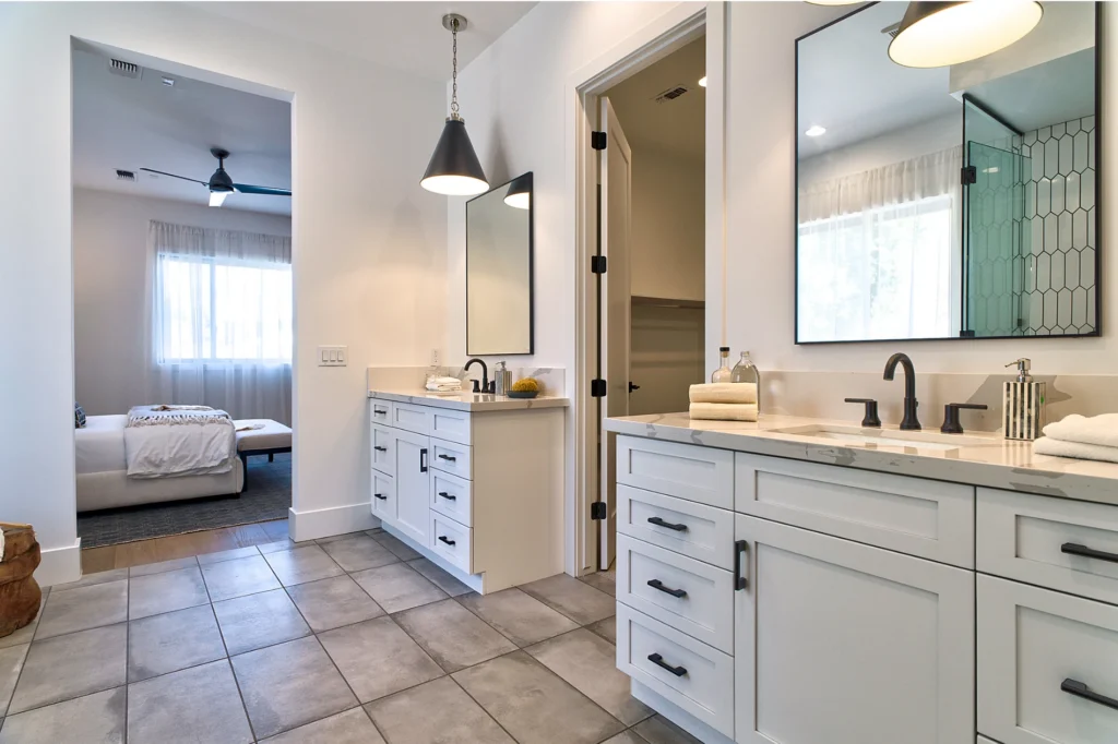 Modern bathroom with dual white vanities, black fixtures, and tile flooring, with a view into an adjacent bedroom.