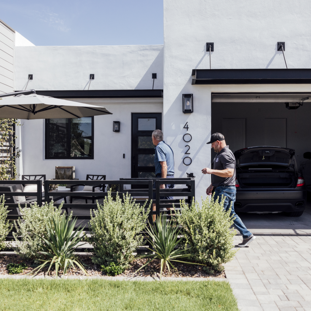 Two men walk into the exterior gate of a modern home with white stucco and black accents