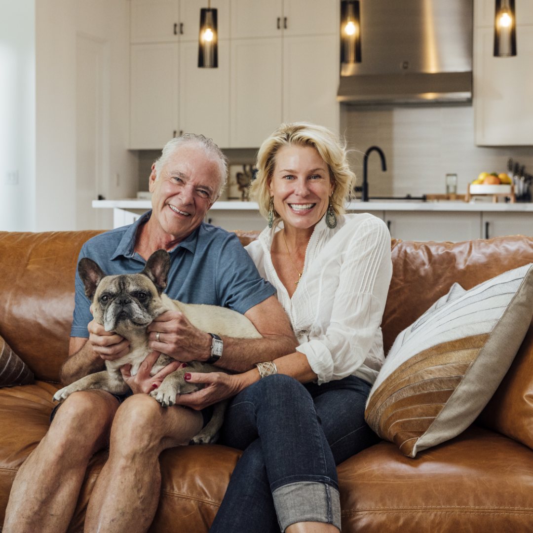 Mike and Robin, TJH happy homeowners pose for a portrait on their brown leather couch with their french bulldog and luxury white kitchen in the background.