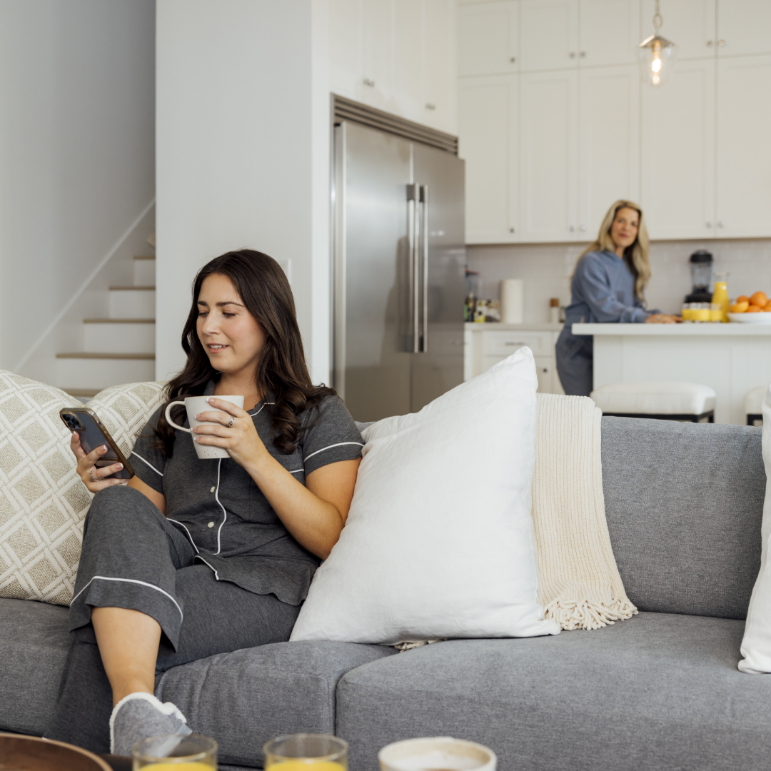 Girl on smartphone drinking coffee with mom in the background preparing breakfast in luxury kitchen