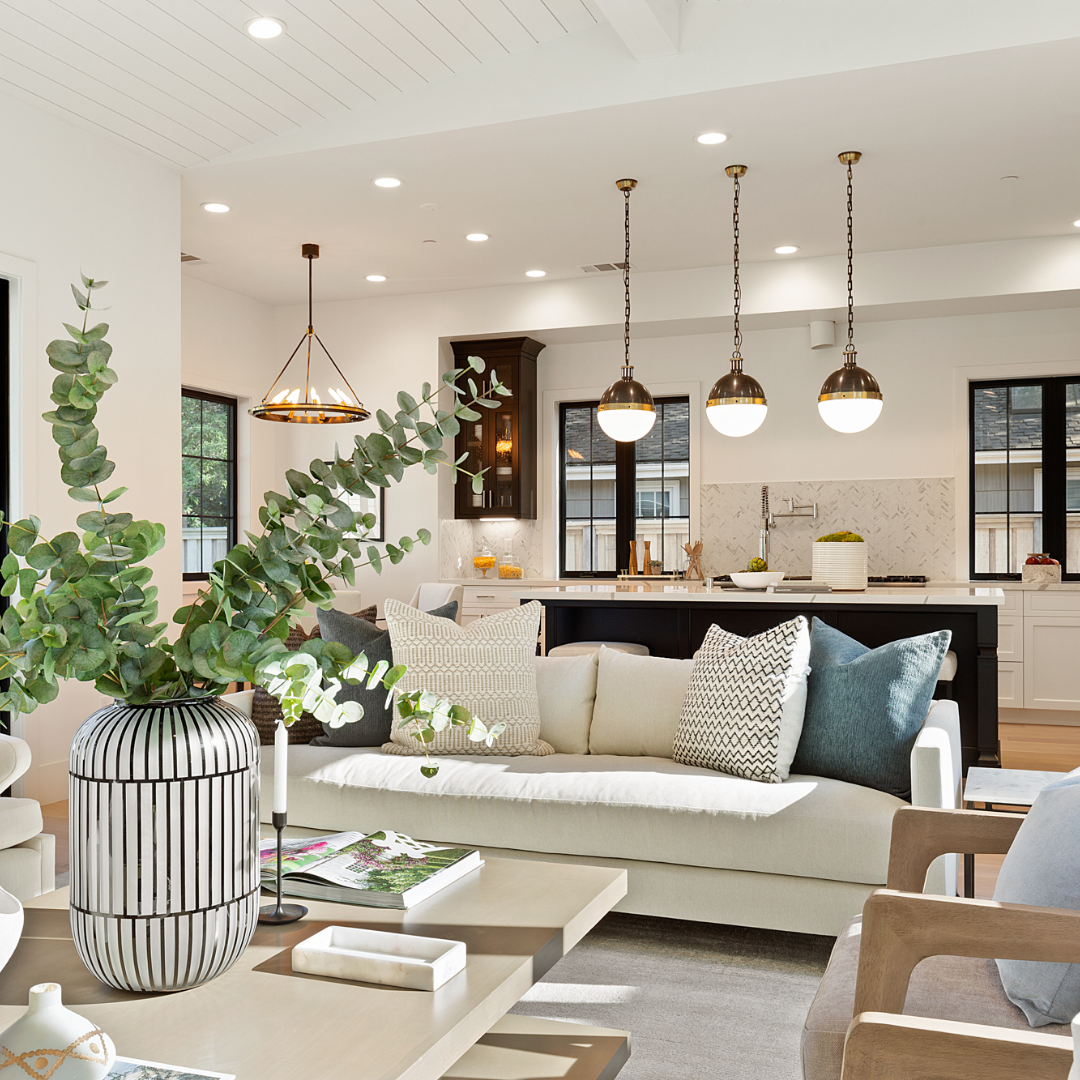 Living room with green plant on coffee table and three hanging lights over kitchen counter with a white sofa and wooden accent chair