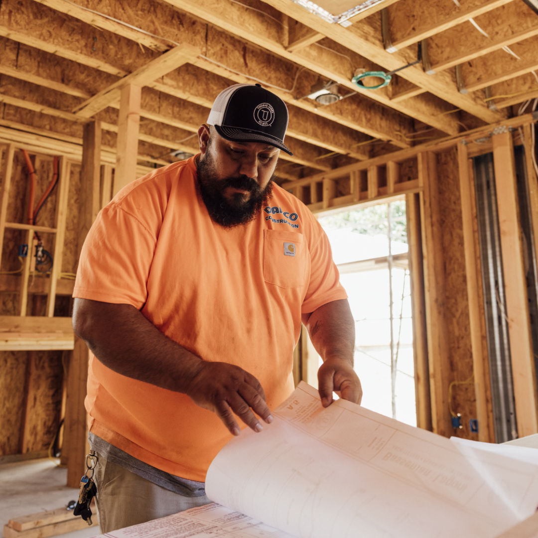 Image of man on construction site wearing an orange shirt and black hat looking through blue prints.