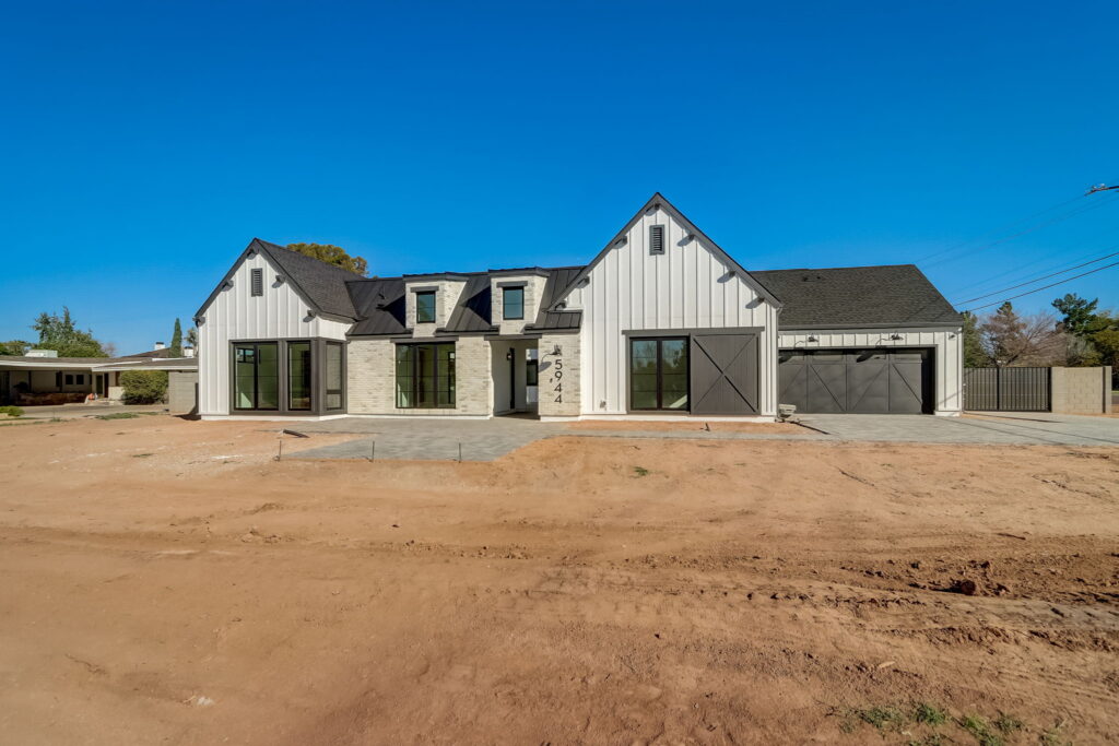Modern farmhouse-style home with light siding, dark roof, and large windows. The front yard is unfinished dirt, and the sky is clear and blue. The house features a garage and gable roofs.