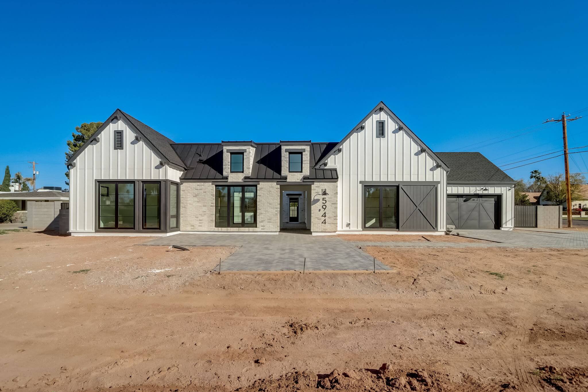 Modern farmhouse-style home with white siding, black-framed windows, a gray roof, and a three-car garage, set on a bare dirt yard under a clear blue sky.
