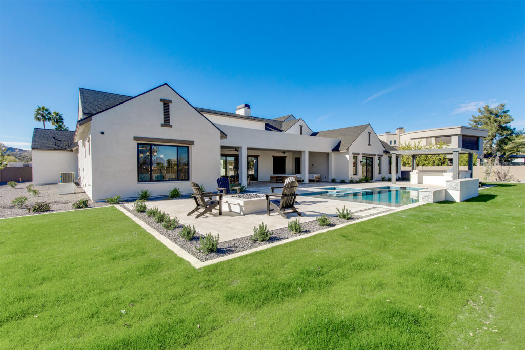 Modern white house with dark roof, large windows, and a spacious backyard featuring a swimming pool, hot tub, fire pit area with chairs, and green lawn under a clear blue sky.