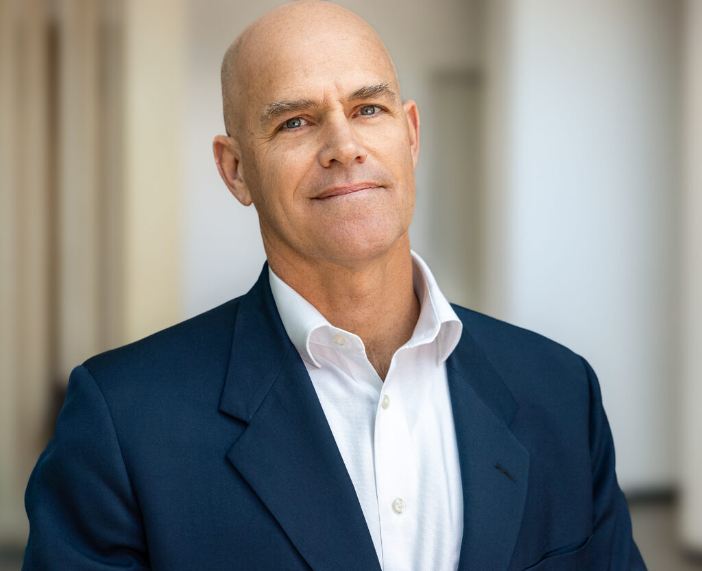 Professional headshot of a man in a navy blazer and white dress shirt, standing indoors with a softly blurred, neutral background.