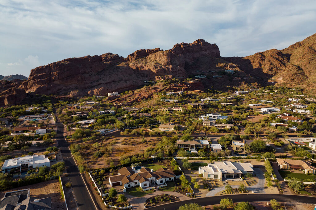 Aerial view of a residential neighborhood with modern homes surrounded by desert landscape, green trees, and rocky mountains in the background under a partly cloudy sky.