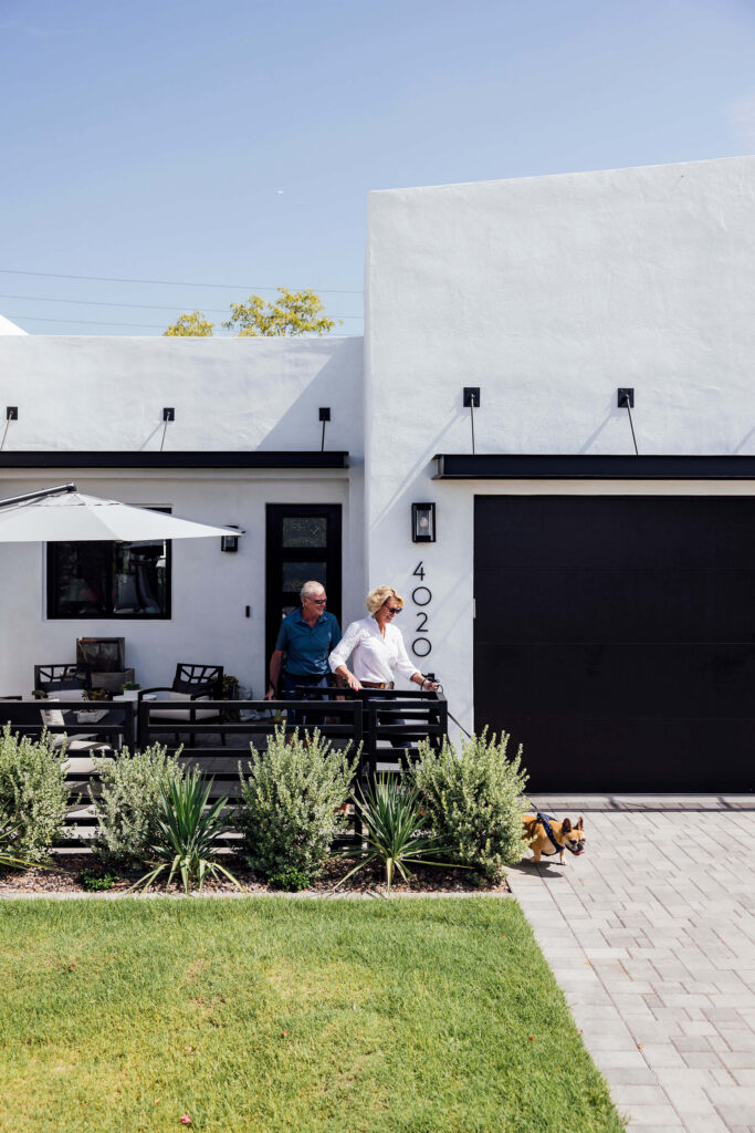 An older couple walks outside a modern white house with black trim and the number 4020 on the wall. They are on a patio near green shrubs, and a small brown dog walks beside them on a paved path.