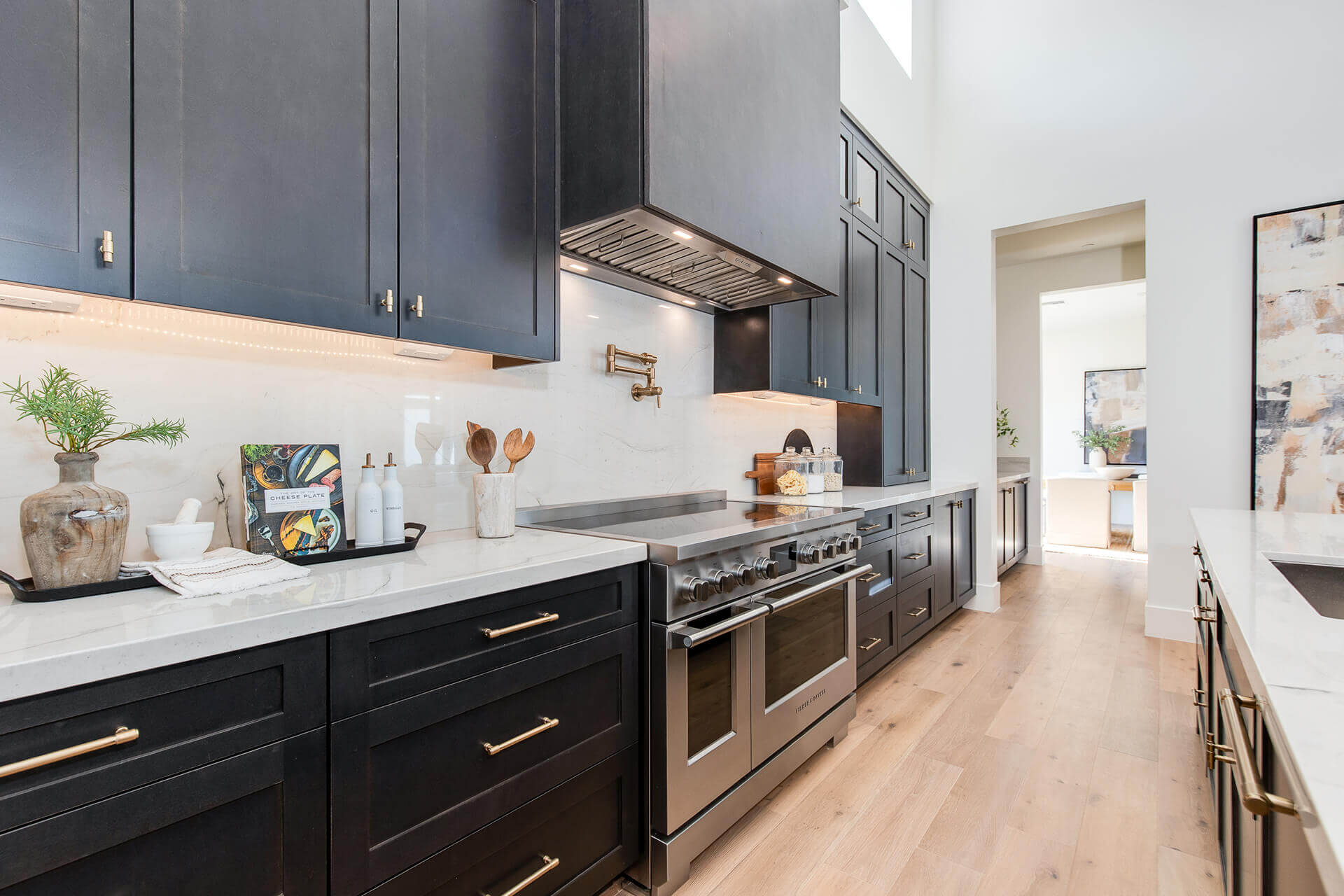 Modern kitchen with dark cabinets, marble countertops, a stainless steel stove with pot filler, light wood floors, and bright natural light. Decorative items and utensils are neatly arranged on the counters.