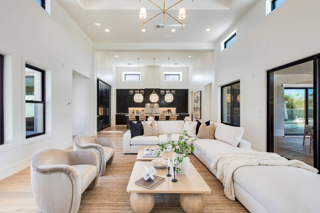 Bright, modern living room with cream sofas, armchairs, a wooden coffee table, large windows, and neutral decor. An open kitchen with pendant lights and a dining island is visible in the background.
