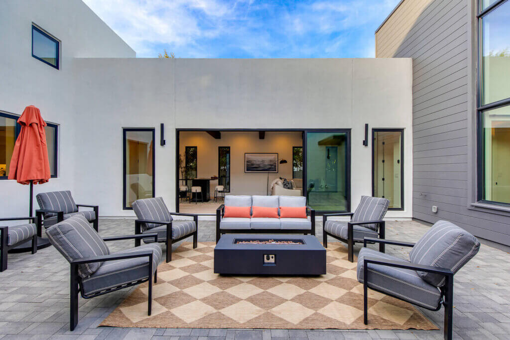 Modern patio with gray cushioned chairs and a sofa arranged around a rectangular fire pit on a patterned rug. The space is surrounded by white and gray walls with large windows and an open door leading inside.