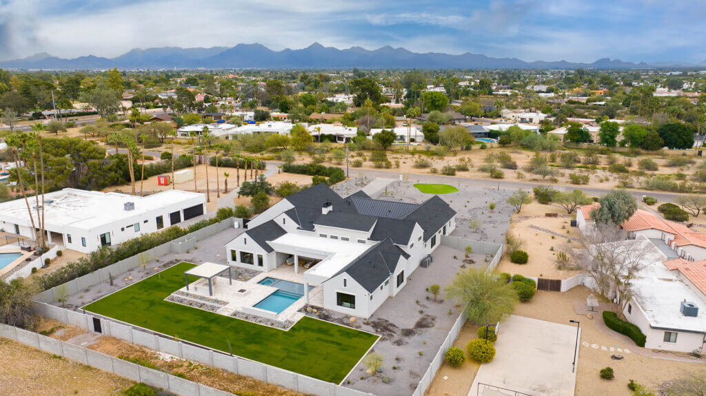 Aerial view of a large modern house with a pool, green lawn, and a covered patio, surrounded by a fenced yard in a suburban neighborhood with mountains in the background.