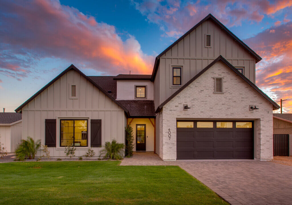 Front exterior of a TJH modern farmhouse‑style home at sunset, featuring light vertical siding, white brick accents, a dark double garage door, warm interior lighting, and a neatly manicured lawn beneath a colorful evening sky.