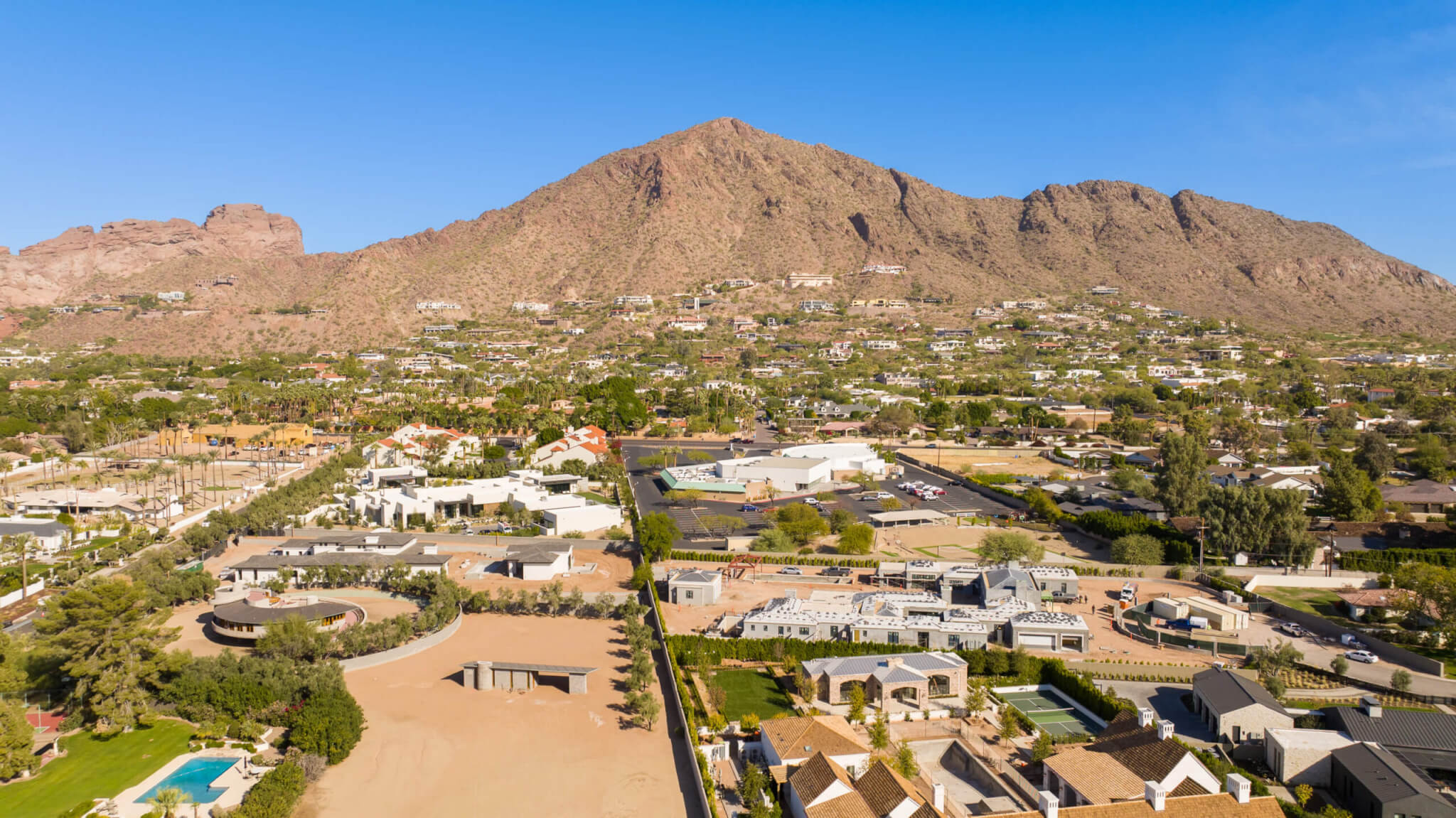 Aerial view of a desert residential neighborhood at the base of a rocky mountain, with luxury homes, tree-lined streets, and clear blue skies in the background.