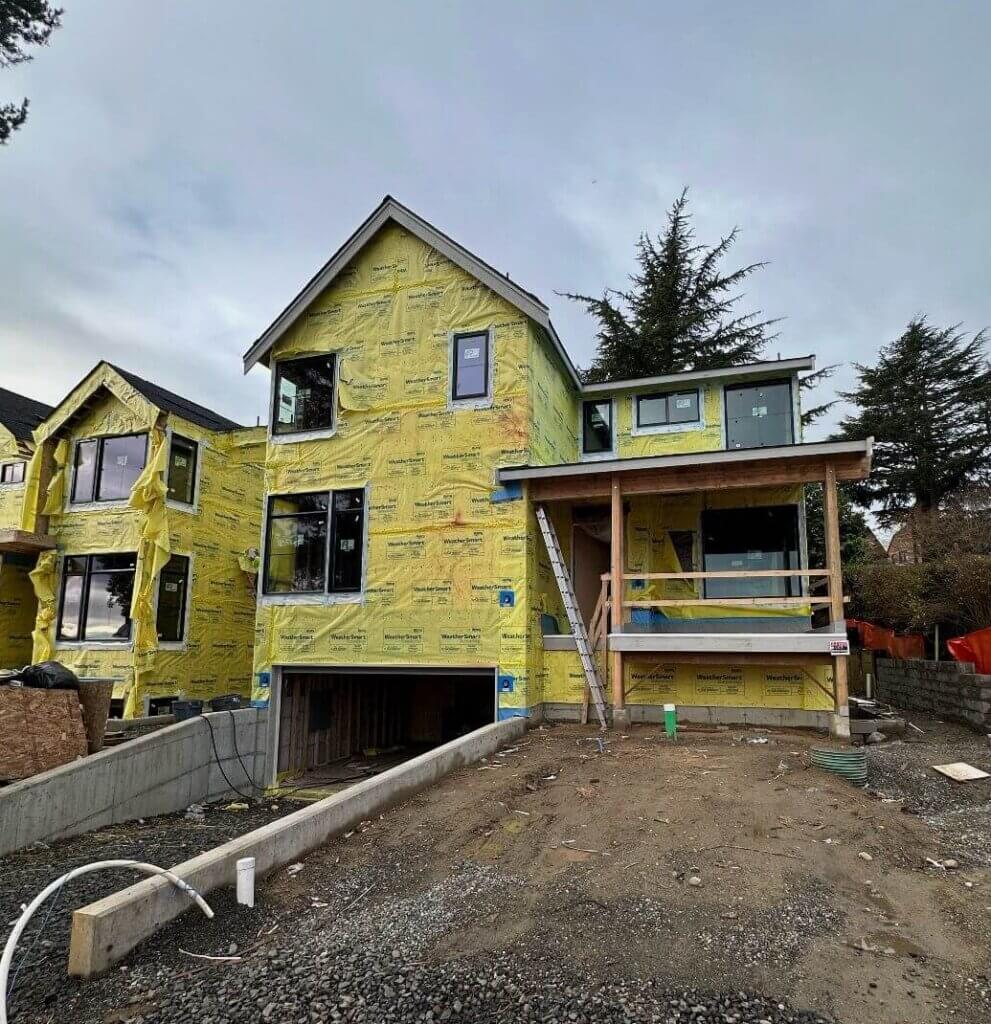 Two-story home under construction with exposed framing and weather barrier, showing early exterior build progress.