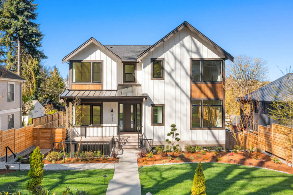 Front exterior of a TJH home with white vertical siding, black-trimmed windows, warm wood accents, a covered porch, and landscaped yard under a clear blue sky.