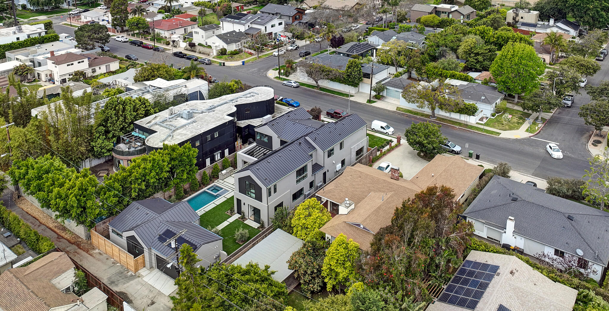 Aerial view of a residential block featuring a contemporary multi‑story home with clean lines and dark roofing, surrounded by traditional single‑family houses, mature trees, and intersecting neighborhood streets.
