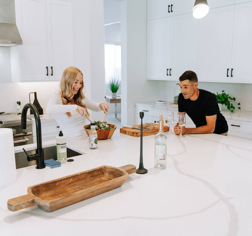 Two people preparing food in a bright, modern white kitchen, standing across a large island with a farmhouse sink, black fixtures, and minimal decor, creating a relaxed, casual cooking moment.