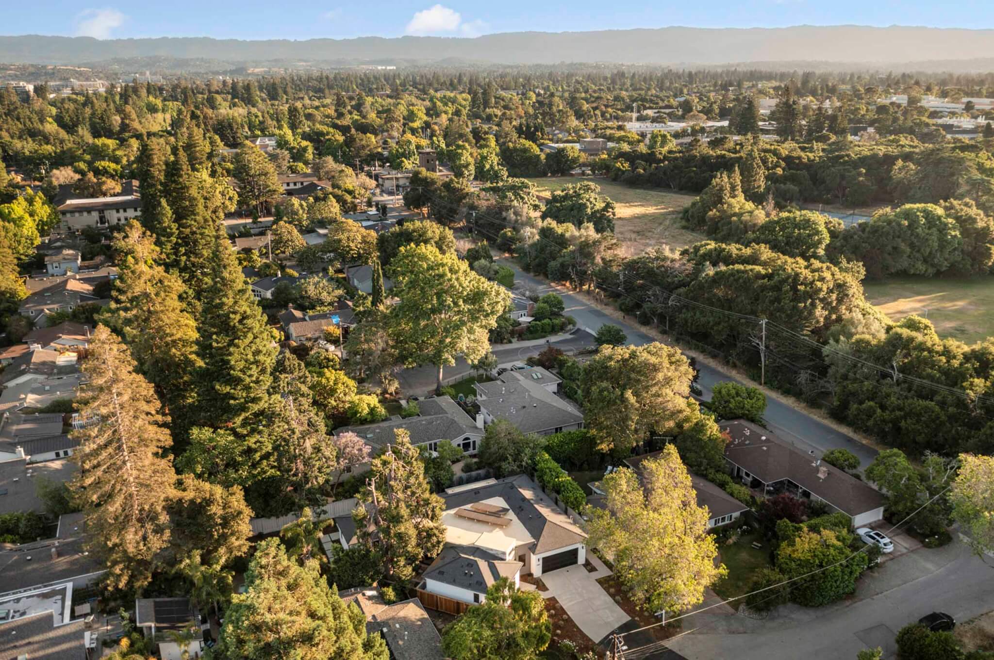 Aerial view of a quiet suburban neighborhood surrounded by mature trees, showing single-family homes, tree-lined streets, and open green spaces with hills in the distance under clear skies.