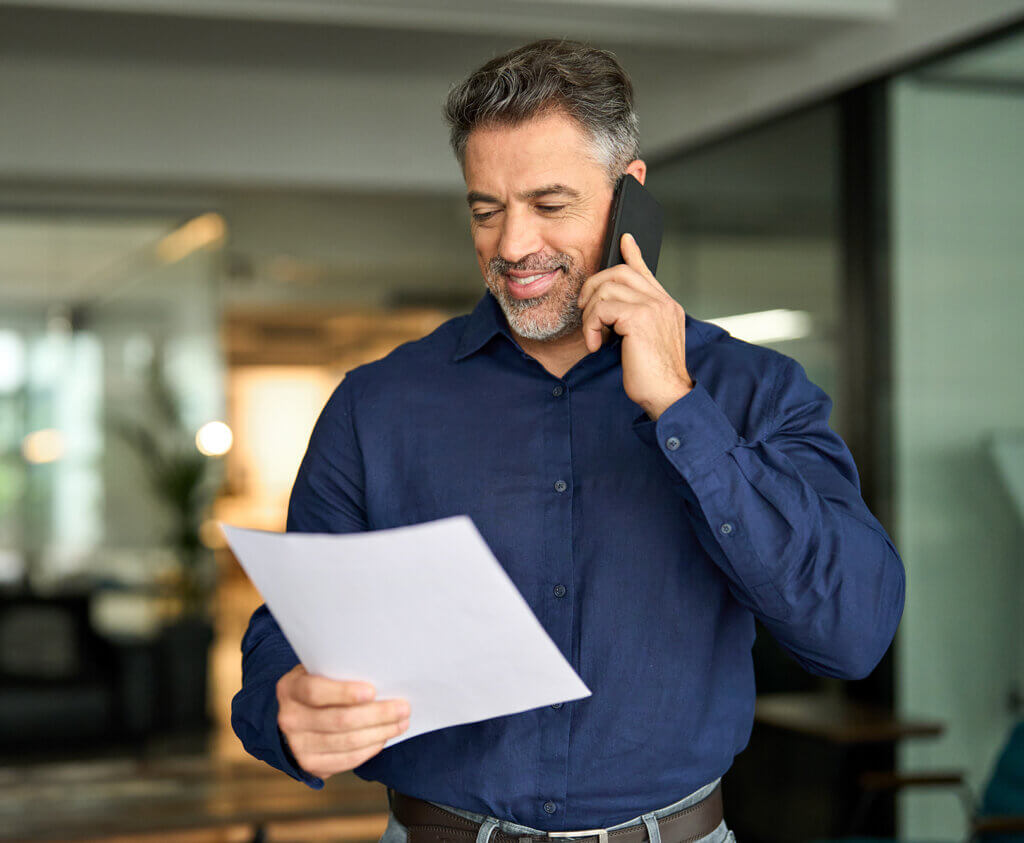 Smiling middle aged senior professional businessman executive looking at documents talking on cell phone standing in office.