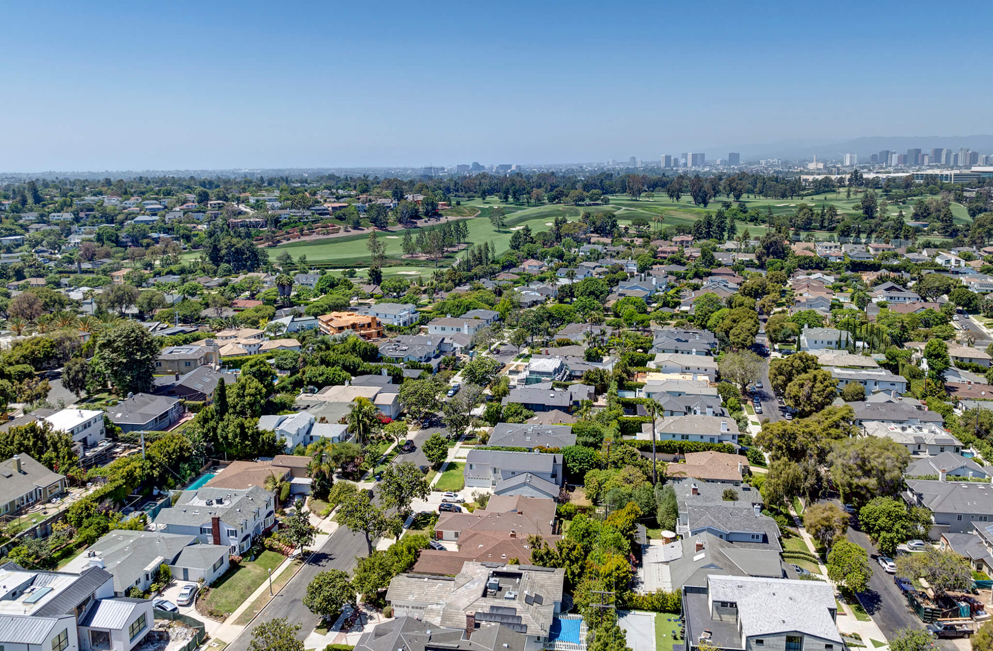 Aerial view of a leafy suburban neighborhood with tightly packed single‑family homes and tree‑lined streets, looking toward a cluster of modern high‑rise buildings on the horizon beneath a clear blue sky.