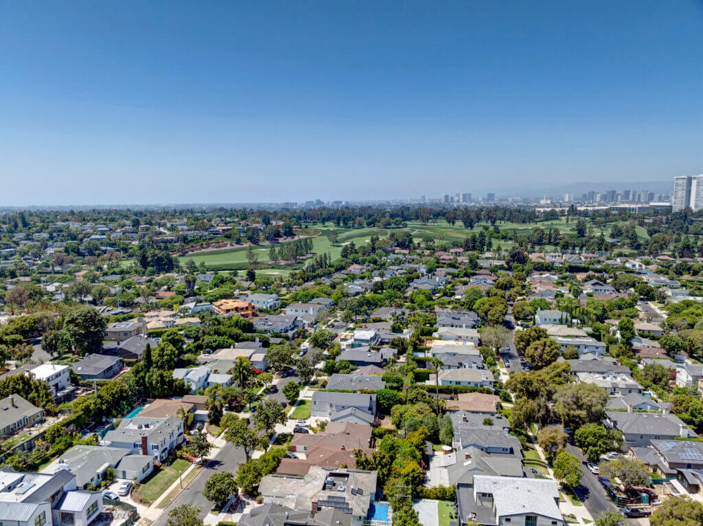 Aerial view of a leafy suburban neighborhood with tightly packed single‑family homes and tree‑lined streets, looking toward a cluster of modern high‑rise buildings on the horizon beneath a clear blue sky.