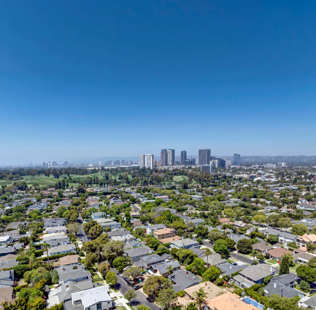 Aerial view of a leafy suburban neighborhood with tightly packed single‑family homes and tree‑lined streets, looking toward a cluster of modern high‑rise buildings on the horizon beneath a clear blue sky.