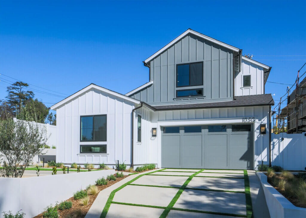 Front exterior of a modern TJH home with gray and white vertical siding, a double garage, large black‑framed windows, and a clean concrete driveway accented with grass strips under a clear blue sky.