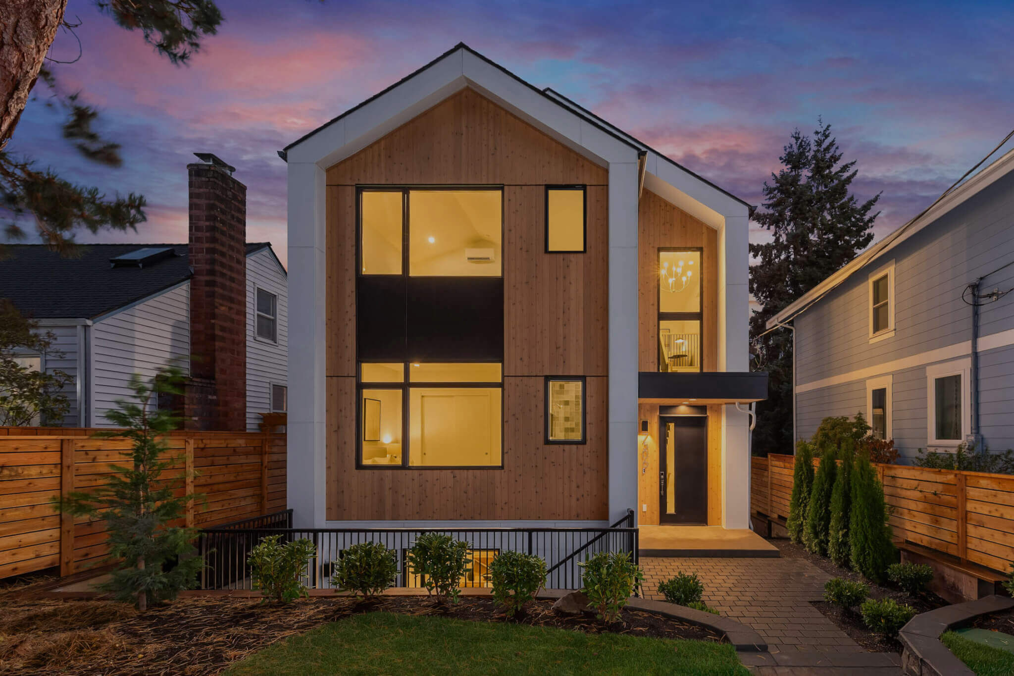 Modern TJH home with a wood-paneled façade, large black-framed windows, and a covered entry, photographed at dusk with warm interior lights and landscaped front yard.