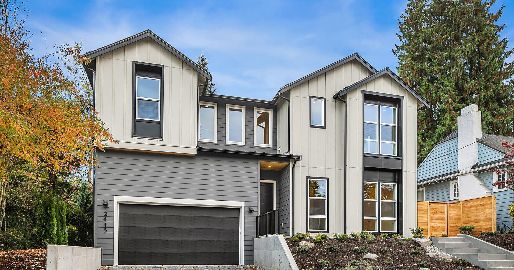 Modern two-story home with mixed siding, large windows, and a garage, set on a landscaped sloped lot.