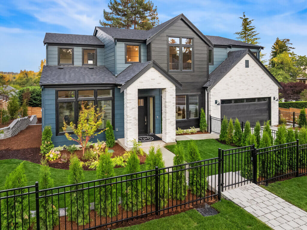 Modern two-story home with gray and blue siding and white brick accents, featuring large black-framed windows, a covered front entry, manicured landscaping, and a black metal fence along the front lawn.