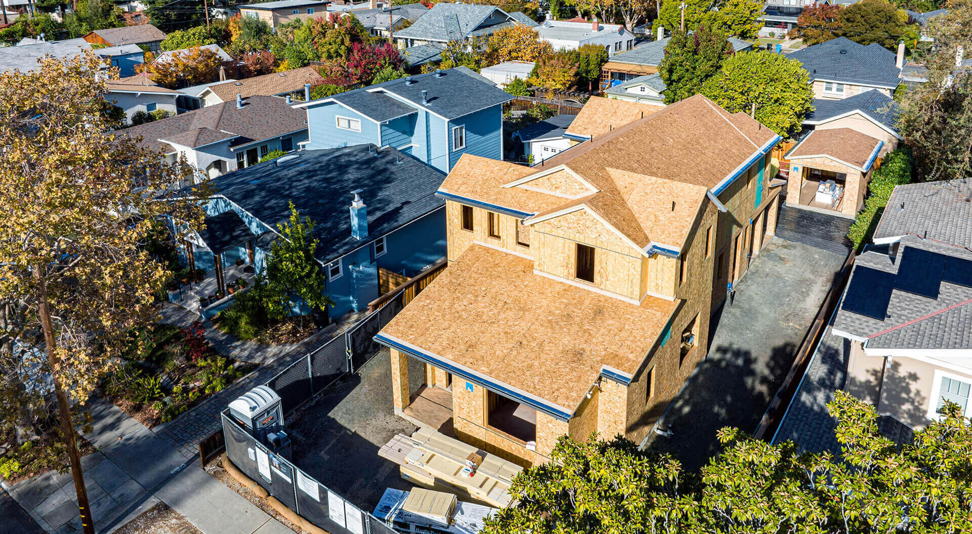 Aerial view of a residential neighborhood with a two-story house under construction in the foreground, surrounded by completed homes, trees with autumn foliage, and clear blue sky.