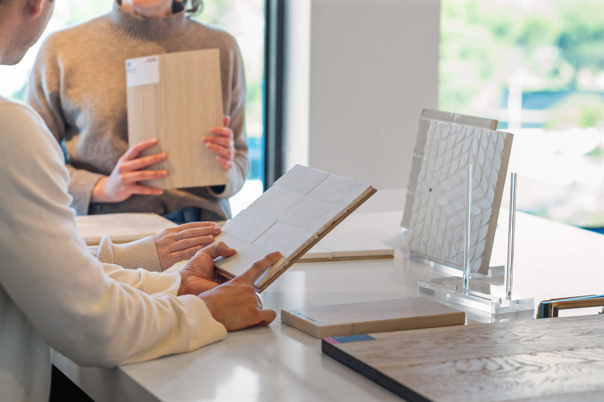 Couple reviewing flooring and tile samples at a TJH Design Studio, including a wood-look plank, white subway tile board, and textured decorative tile display