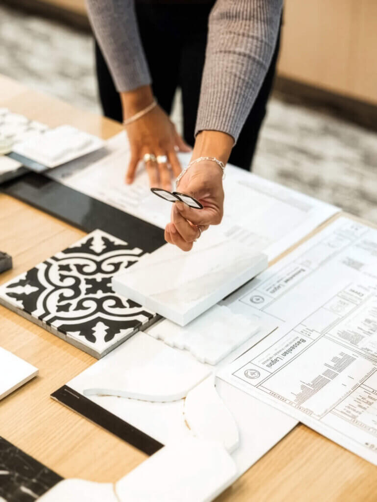 TJH Design Studio consultant reviewing tile and material selections, including a decorative black and white encaustic tile, marble samples, and architectural floor plans