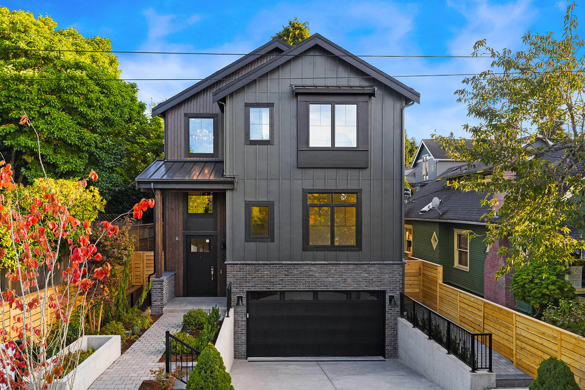Modern three-story home with dark siding, large windows, and a front entry above a garage, surrounded by trees and landscaping.