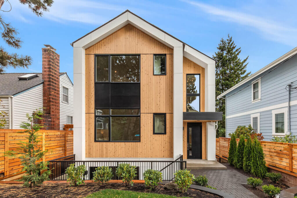 A modern two-story house with light wood siding, large black-framed windows, and white trim. The front yard has young landscaping, a paved walkway, and is enclosed by a wooden fence. Neighboring houses are visible on both sides.