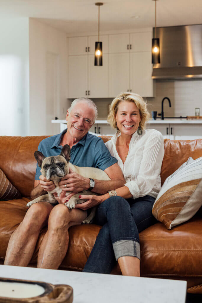 Smiling couple relaxing on a cognac leather sofa with their French Bulldog in a TJH home featuring an open-concept layout