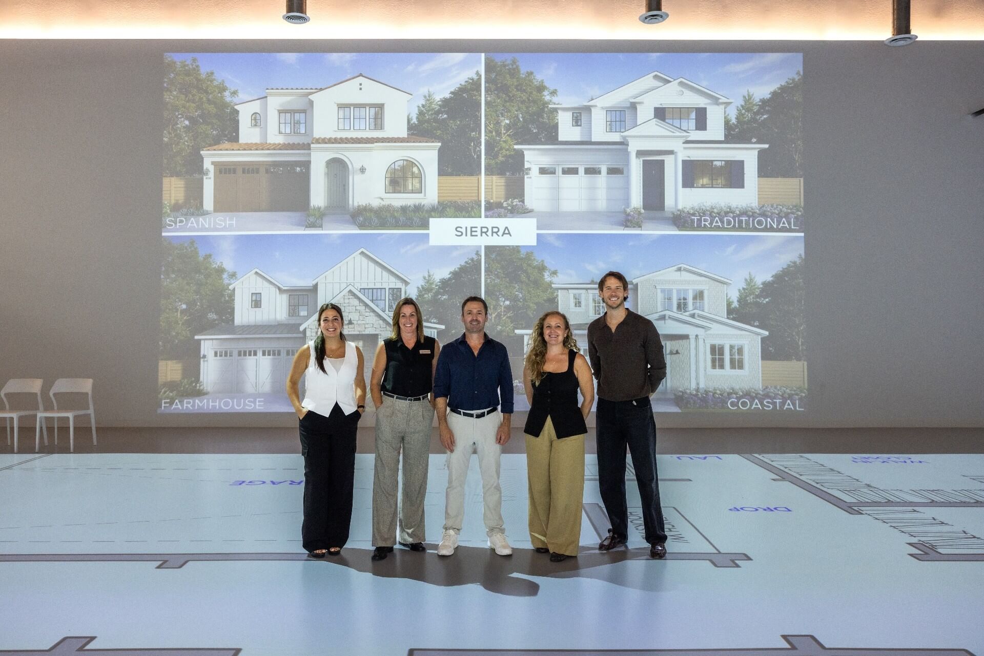 Five people stand on a projected floor plan in front of Sierra home renderings in Spanish, Traditional, Farmhouse and Coastal architectural styles.